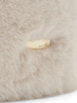 Close-up of a soft, fluffy cream-colored faux fur or sheepskin material with a small rectangular gold-tone metal tag embossed with the brand name 'SNIDEL'.