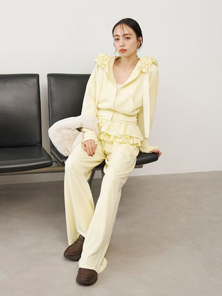 A model sits on a black bench wearing a pale yellow ruffled two-piece set with wide-leg pants and brown loafers, against a minimalist gray wall.