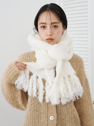 A woman wearing a thick, fluffy off-white scarf with fringe details and a beige knit cardigan, standing against a white wall with a louvered door in the background.