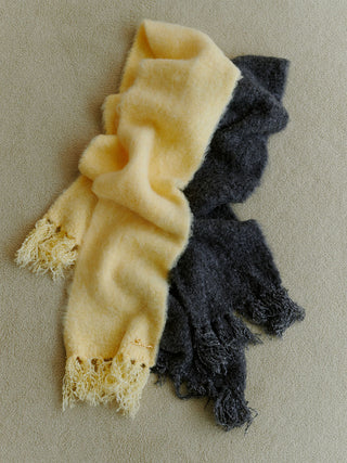 Two fuzzy fringed scarves laid flat on a neutral beige background, one in pale butter yellow and the other in dark charcoal gray.