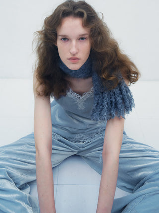 A model with wavy light brown hair sits on a white floor wearing a dusty blue velvet dress with lace trim, a matching blue knit scarf, and a fringed blue shawl.