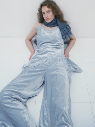 A model wearing a light blue velvet jumpsuit with lace trim and a matching blue knit scarf, lying on a white tiled floor against a white wall.