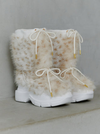A pair of white platform boots with fluffy faux-fur panels in a beige and white spotted pattern, featuring white laces with gold tips, set against a gray concrete background.