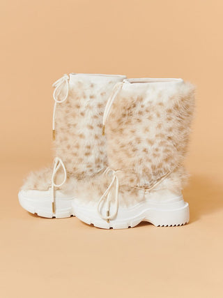 A pair of white snow boots with fluffy beige faux fur shafts and leopard-like spots, featuring white laces and chunky white soles, set against a solid peach background.