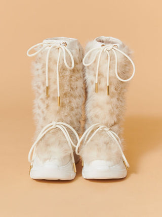 A pair of tall, fluffy faux-fur boots in a light beige and white pattern, featuring white laces with gold tips and white soles, set against a solid peach background.