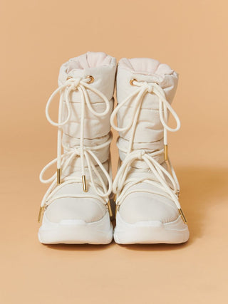A pair of off-white high-top lace-up boots with gold-tone hardware, shown from the front against a solid peach background.
