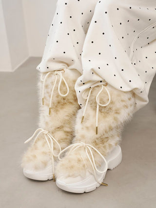 A close-up of a person wearing fluffy cream faux-fur lace-up boots with white platform soles and white drawstring hems, paired with white pants featuring black polka dots.