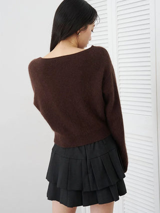 A woman wearing a dark brown off-the-shoulder knit sweater and a black pleated mini skirt, photographed from behind against a white paneled wall.