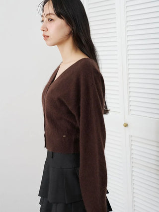 A woman in profile wearing a dark brown knit cardigan and a black pleated skirt, standing in front of white louvered doors.
