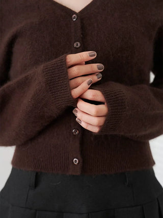 A close-up of a person wearing a dark brown fuzzy knit cardigan with button-front closure and ribbed cuffs, paired with black bottoms. The person's hands with dark gray manicured nails are clasped in front.
