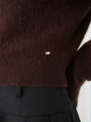Close-up of a dark brown fuzzy knit sweater with a ribbed hem, featuring a small oval gold button detail, paired with dark gray pleated trousers.