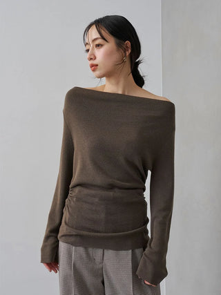 A woman wearing a dark brown off-the-shoulder knit sweater and gray patterned trousers, standing against a light gray wall.