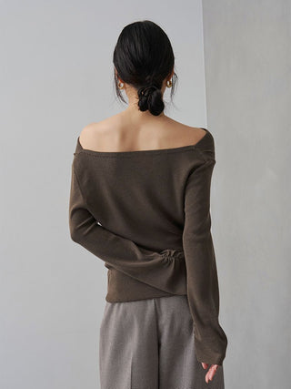 A woman shown from the back wearing a dark brown off-the-shoulder knit top and textured taupe pants against a minimalist gray background.