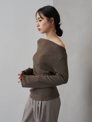 A woman wearing a brown off-the-shoulder knit sweater and gray striped trousers, standing in profile against a plain light gray wall.