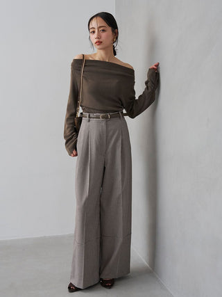A woman stands against a light gray wall wearing a dark brown off-the-shoulder long-sleeve top and high-waisted, wide-leg taupe trousers with a thin belt. She is carrying a small beige shoulder bag.