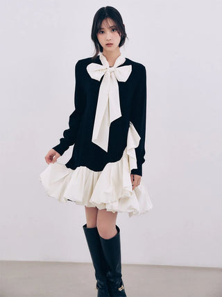 A model wearing a black knit sweater with a large white bow at the collar, layered over a white ruffled mini skirt, and paired with black knee-high boots, set against a plain light gray studio background.