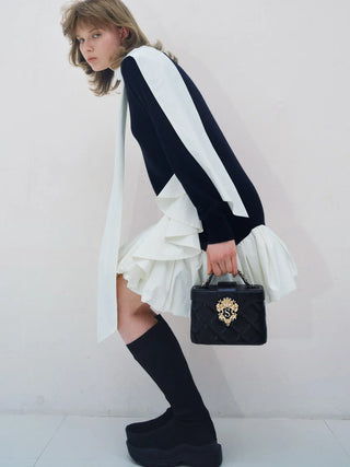 A model wearing a black and white color-blocked long-sleeve top with a white ruffled hem, black over-the-knee socks, and holding a black quilted handbag with a gold crest.