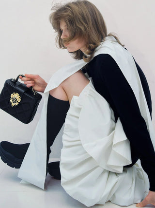 A model in a black and white outfit kneels on a white floor, holding a black quilted handbag with a gold crest logo. The outfit features a dramatic white draped vest over a black long-sleeve top and black thigh-high socks.