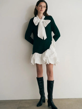 A model stands against a plain off-white wall wearing a dark green long-sleeve sweater with a large white bow at the collar, layered over a white ruffled mini skirt, and wearing black knee-high leather boots.