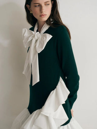 A model wearing a dark green knit sweater layered over a white ruffled blouse with a large white bow at the collar, photographed against a plain light wall.