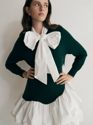 A woman wearing a dark green long-sleeve sweater with a large white bow at the collar and a white ruffled skirt, posing against a plain light background.