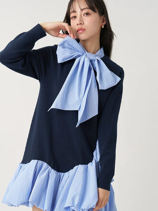 A model wearing a navy blue long-sleeve knit top with a light blue and white striped bow tie-neck detail and a matching striped ruffled hem.