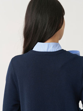 A woman shown from the back wearing a navy knit sweater over a light blue and white striped collared shirt, against a plain light background.