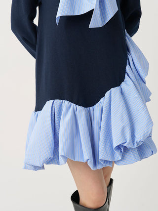 A close-up of a model wearing a long-sleeve dress with a solid navy blue upper section and a light blue and white striped ruffled hem, paired with black boots.