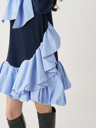 A close-up of a person wearing a navy dress with layered light blue and white striped ruffle trim and a matching striped bow at the neckline, paired with black knee-high boots against a plain light background.