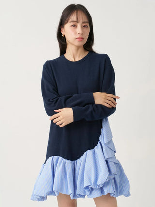 A woman modeling a long-sleeve knit top with a dark navy upper section and a light blue and white vertically striped ruffled hem, against a plain light gray studio background.