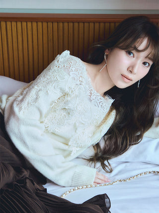 A woman reclining on a bed wearing a cream knit sweater with intricate lace applique on the chest and sleeves, paired with a dark brown pleated skirt. She is styled in a bedroom setting with a wooden headboard and white bedding.