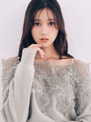 A woman wearing a light gray knit sweater with a sheer lace overlay across the chest, posed against a plain light background.