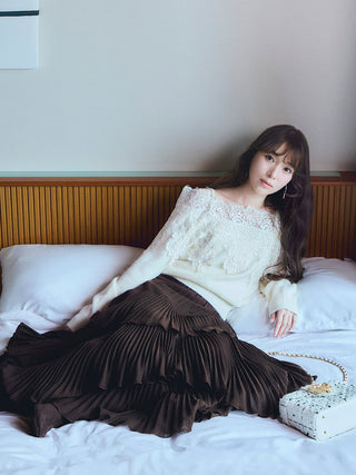 A woman reclining on a bed wearing a cream lace-trim off-the-shoulder sweater and a dark brown pleated tiered skirt, with a small white patterned handbag beside her.