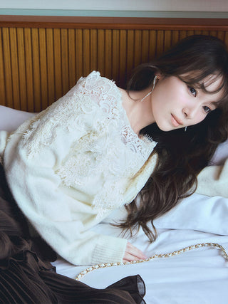 A woman reclining on a bed wearing a cream knit sweater with intricate lace applique on the shoulders, styled with a dark pleated skirt and a gold chain accessory.