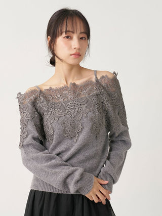 A woman wearing a gray off-the-shoulder knit sweater with intricate lace detailing across the chest and shoulders, paired with a black skirt, against a plain light gray background.