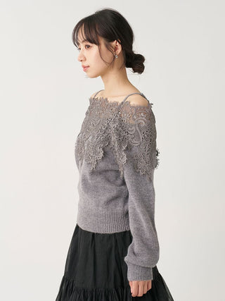 A woman wearing a gray knit sweater with a lace-embroidered off-the-shoulder yoke, paired with a black skirt, photographed against a plain light gray studio background.