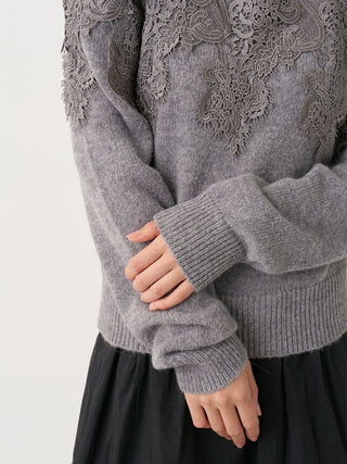 A close-up of a person wearing a gray knit sweater with intricate lace detailing across the chest and shoulders, paired with a black skirt, against a plain light background.