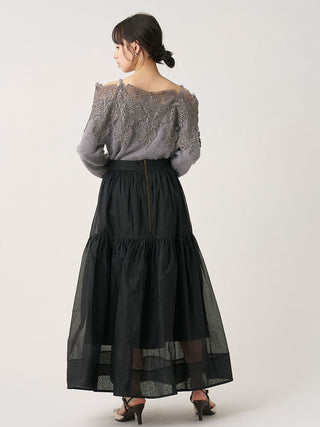 A woman shown from the back wearing a gray lace off-the-shoulder top and a long black tiered skirt with sheer mesh layers, photographed against a plain light gray studio background.
