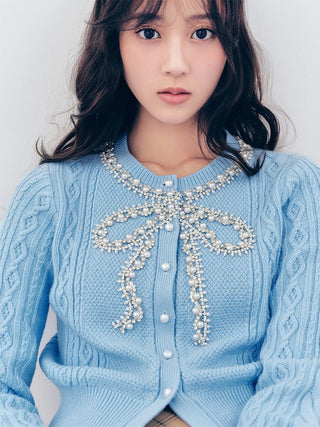 A close-up of a woman wearing a light blue cable-knit cardigan sweater with a large pearl-beaded bow detail across the chest, set against a plain light gray background.