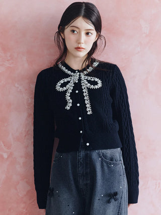 A woman wearing a black cable-knit cardigan with a white floral bow embellishment at the collar, paired with dark gray jeans, posed against a textured pink wall.