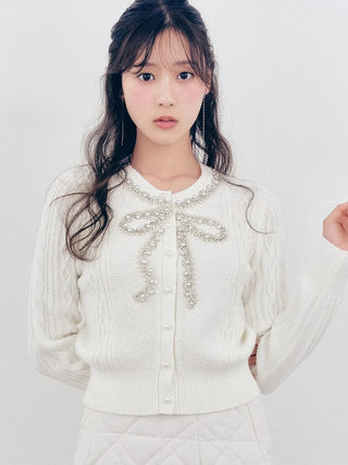 A young woman with long dark hair wearing a cream-colored cable-knit cardigan sweater with a decorative pearl bow detail at the collar, paired with a matching quilted skirt against a plain light background.
