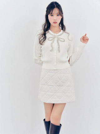 A young woman modeling a white knit cardigan with a pearl-embellished bow at the collar and a matching white quilted mini skirt, paired with black knee-high boots, against a plain white studio background.