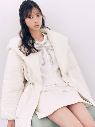 A woman wearing a cream quilted puffer jacket with a faux fur collar, a matching cream knit cardigan with a beaded bow, and a cream mini skirt, seated against a plain light background.