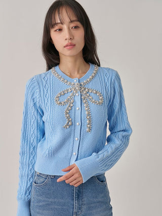 A woman wearing a light blue cable-knit cardigan sweater with a large pearl-beaded bow detail at the neckline, paired with light-wash blue jeans, against a plain light gray studio background.