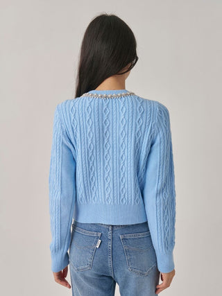 A woman viewed from the back wearing a light blue cable-knit sweater with a beaded collar and blue jeans, set against a plain light gray studio background.