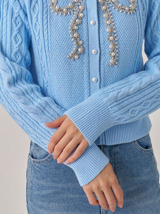 A close-up of a person wearing a light blue cable-knit cardigan with pearl and crystal embellishments on the chest, paired with blue jeans.