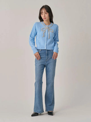 A woman stands against a light gray studio background wearing a light blue long-sleeve button-front top with pearl embellishments at the neckline, paired with light blue flared jeans and black pointed shoes.