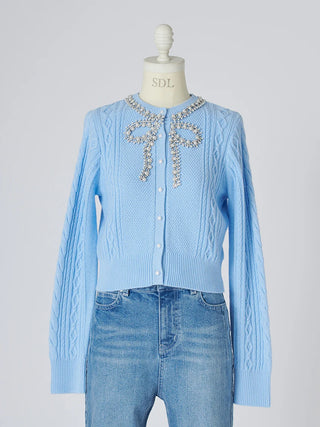 A light blue cable-knit cardigan sweater displayed on a cream dress form mannequin, styled with blue jeans. The cardigan features a decorative silver rhinestone bow detail at the collar and white buttons down the front.
