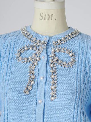 A light blue cable-knit cardigan sweater displayed on a cream mannequin torso. The cardigan features a round neckline adorned with elaborate silver-tone beaded and pearl bow embellishments, with white buttons down the front.