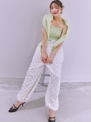 A woman models a light green knit shrug draped over her shoulders, paired with a light green camisole, white drawstring pants with black polka dots, and black kitten-heel mules, standing next to a metal stool against a light gray wall.